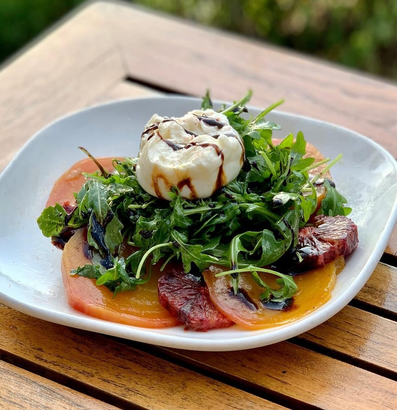 A burrata salad with arugula, sliced tomatoes, and balsamic glaze on a white plate.